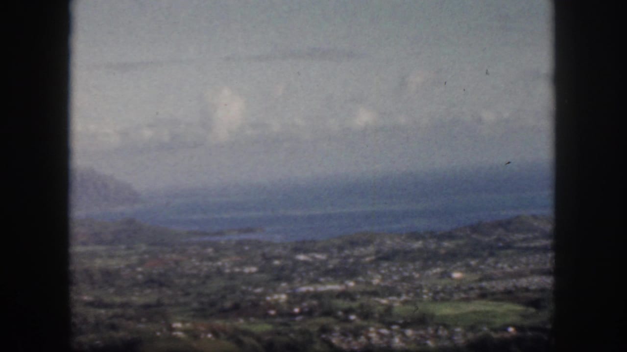1959 Oahu aerial frame with dark aircraft window edges around a soft view of coastal water, town areas, green land, and distant ridges.