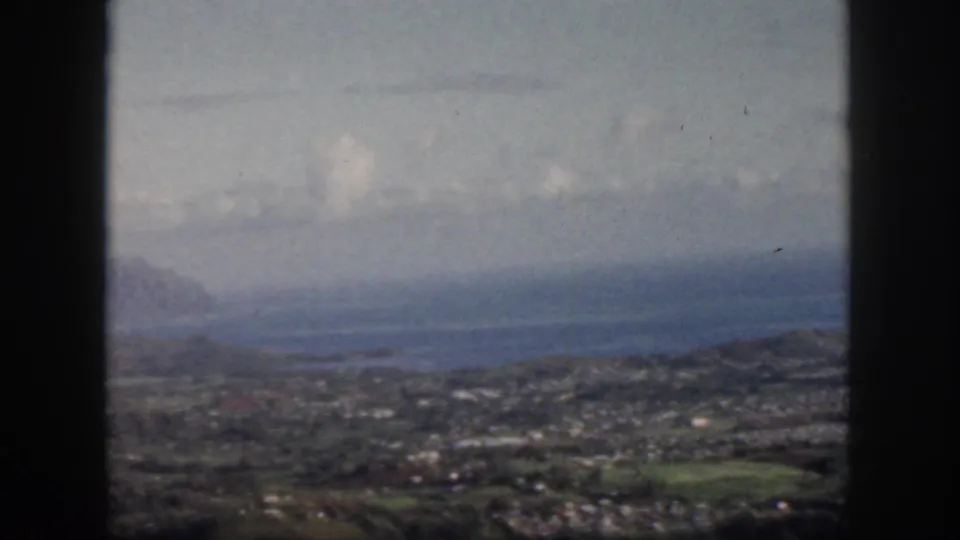 1959 Oahu aerial frame with dark aircraft window edges around a soft view of coastal water, town areas, green land, and distant ridges.