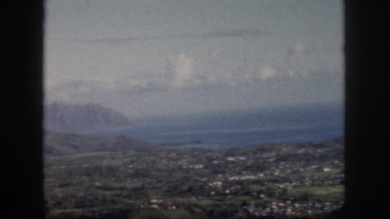 Distant mountains, blue water, low hills, and town structures seen from an airplane window.