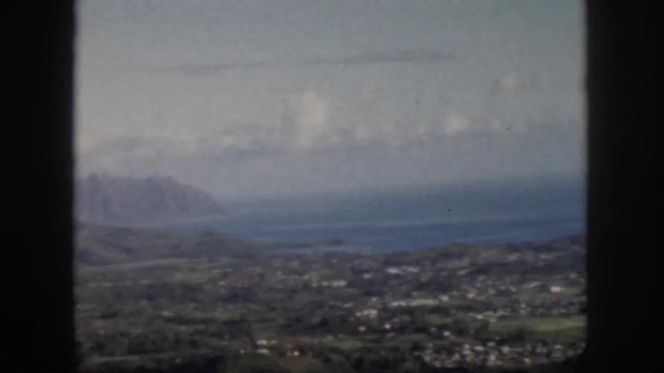 1959 Oahu home-movie still showing a blurred airplane-window view across green lowlands toward blue water and a distant mountain range.