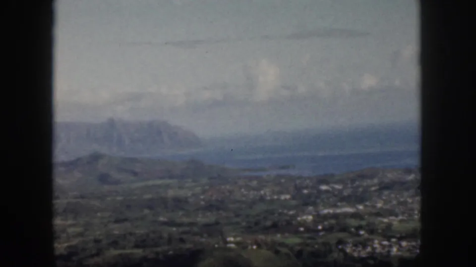 1959 Oahu frame with a dark airplane-window border around a hazy aerial view of settlement, green terrain, coastal water, and mountains.