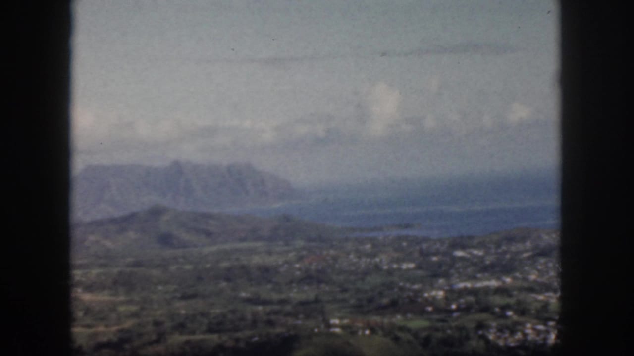 1959 Oahu home-movie frame showing green hills, distant mountains, sky, and dark window mullions in the foreground.