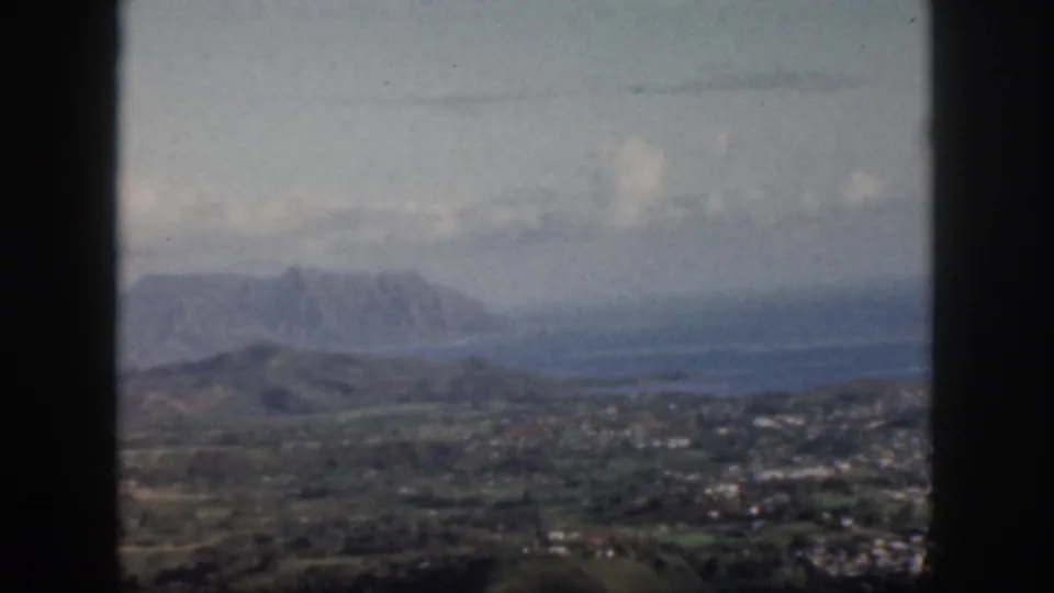 1959 Oahu home-movie frame showing green hills, distant mountains, sky, and dark window mullions in the foreground.