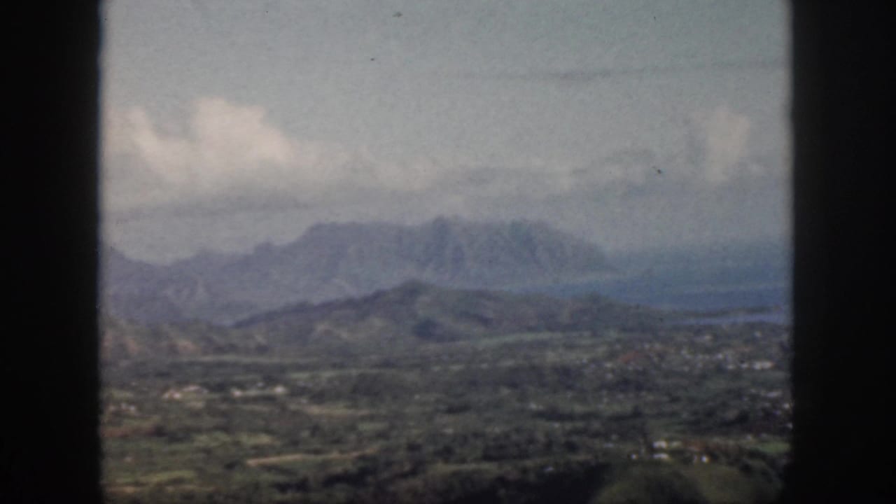 1959 Oahu home-movie still with green hills, distant mountains, bright clouds, and a dark window edge in the foreground.