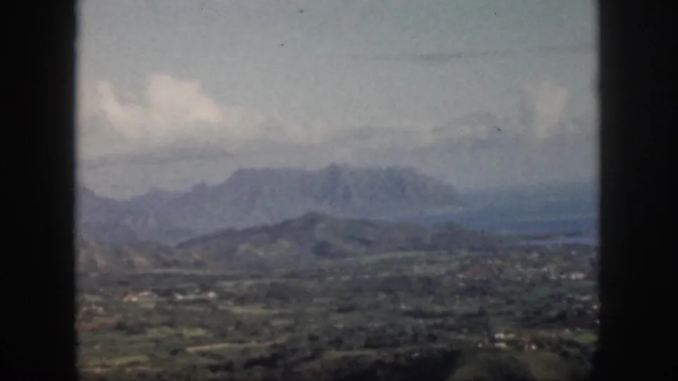 1959 Oahu home-movie still with green hills, distant mountains, bright clouds, and a dark window edge in the foreground.