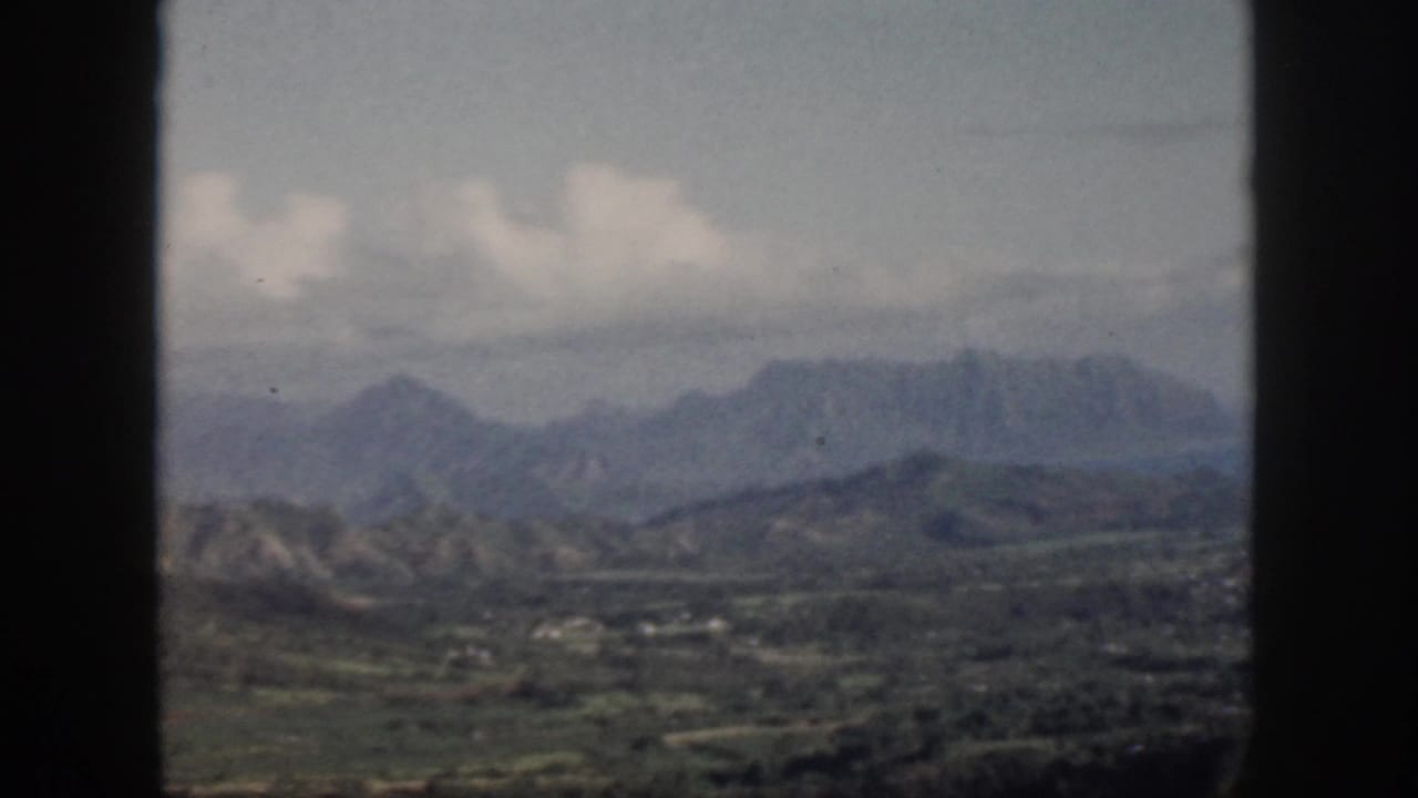 1959 Oahu home-movie frame showing hazy mountain ridges, green valley, clouds, and dark vertical window bars.