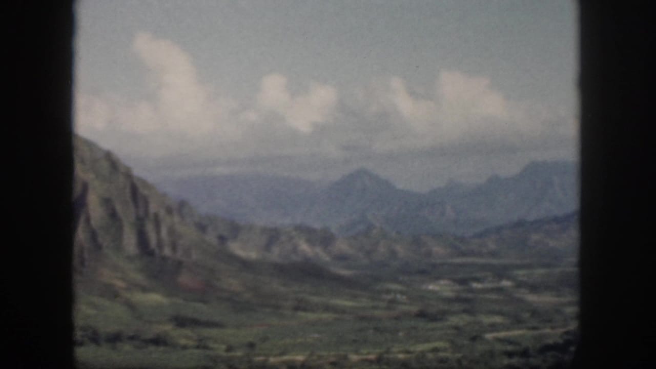 1959 Oahu frame with a steep rocky slope at left, distant mountains, valley floor, and soft clouds through a dark window frame.