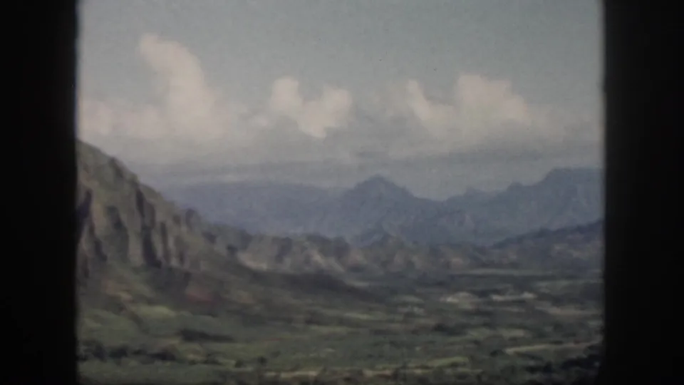 1959 Oahu frame with a steep rocky slope at left, distant mountains, valley floor, and soft clouds through a dark window frame.