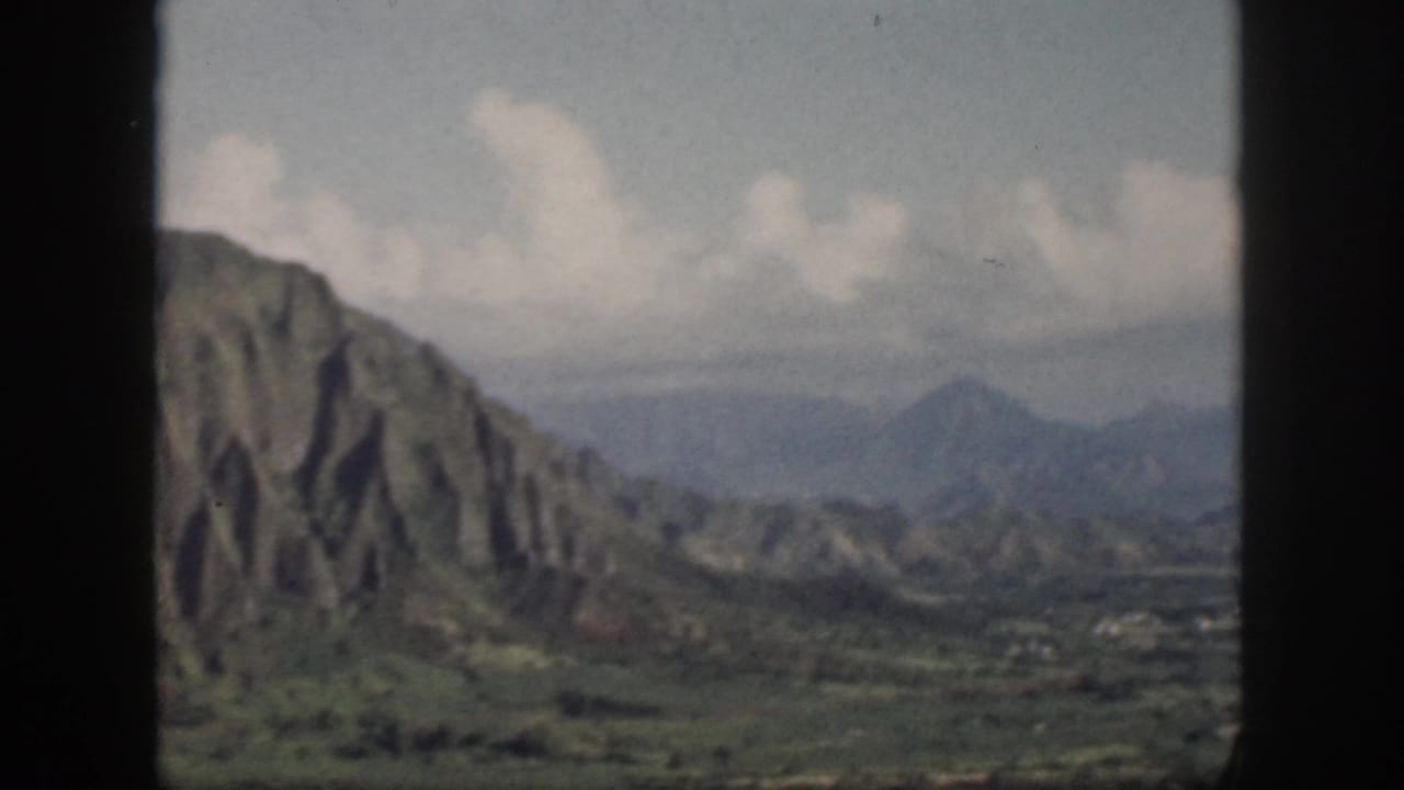 1959 Oahu home-movie still showing a steep ridged mountain face, green lowland, distant peaks, and clouds through window bars.