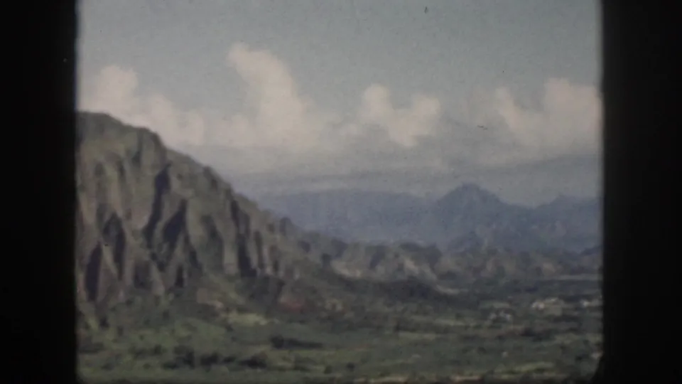 1959 Oahu home-movie still showing a steep ridged mountain face, green lowland, distant peaks, and clouds through window bars.