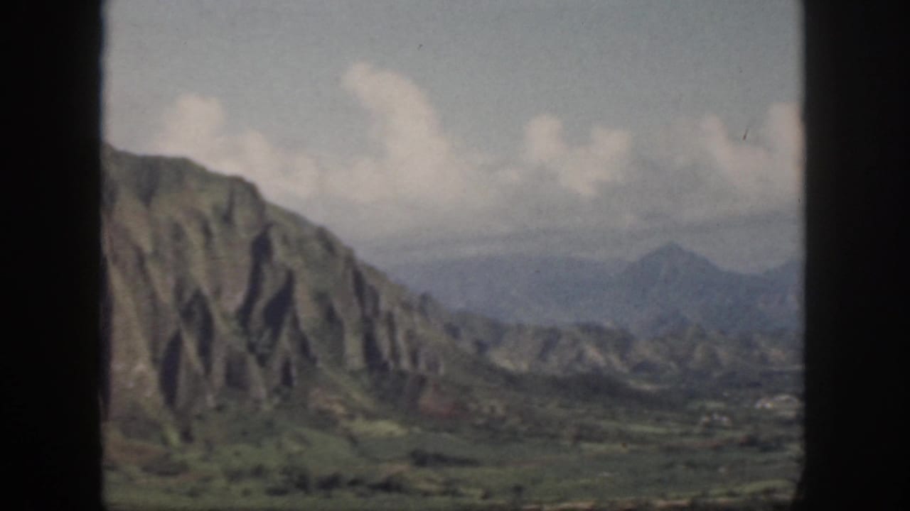 1959 Oahu 8mm frame showing a steep green ridge, hazy valley, distant peaks, and pale clouds.