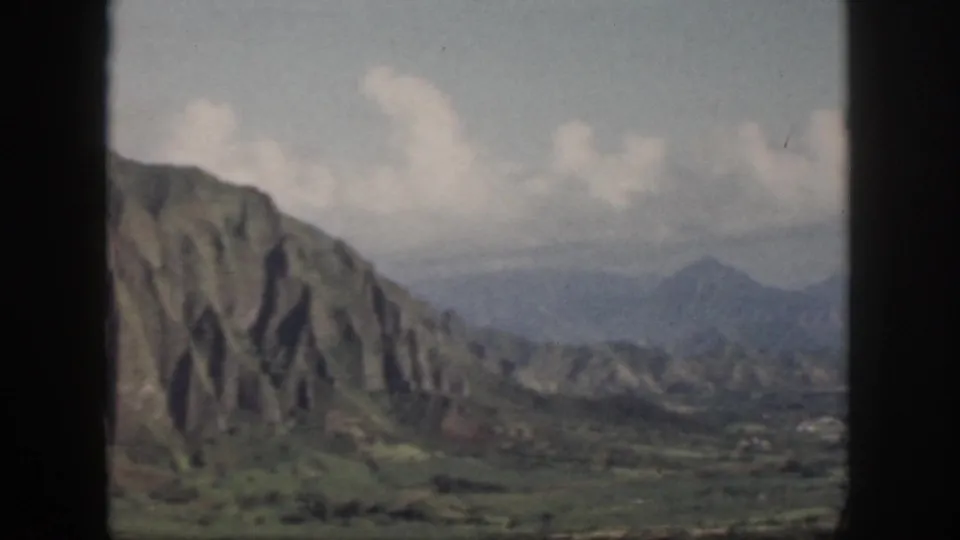 1959 Oahu 8mm frame showing a steep green ridge, hazy valley, distant peaks, and pale clouds.