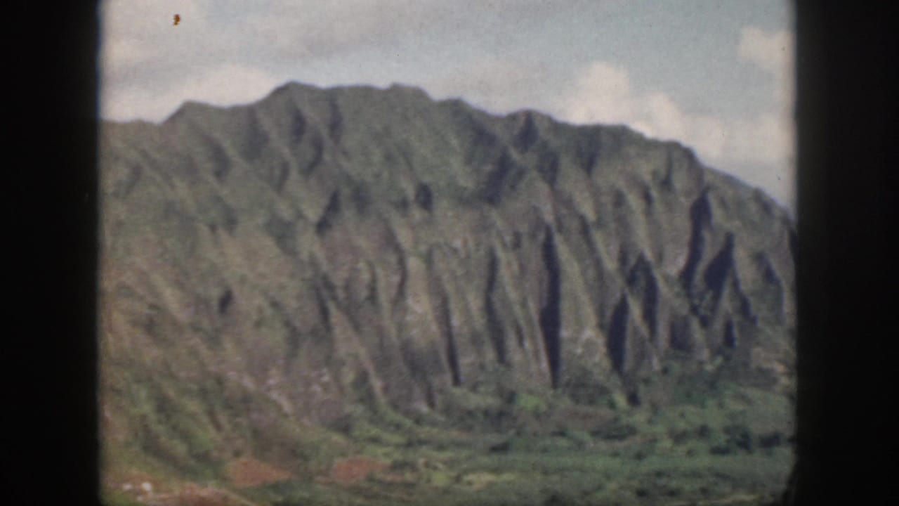 1959 Oahu 8mm frame of a wide green mountain wall with eroded vertical ridges and low valley vegetation.