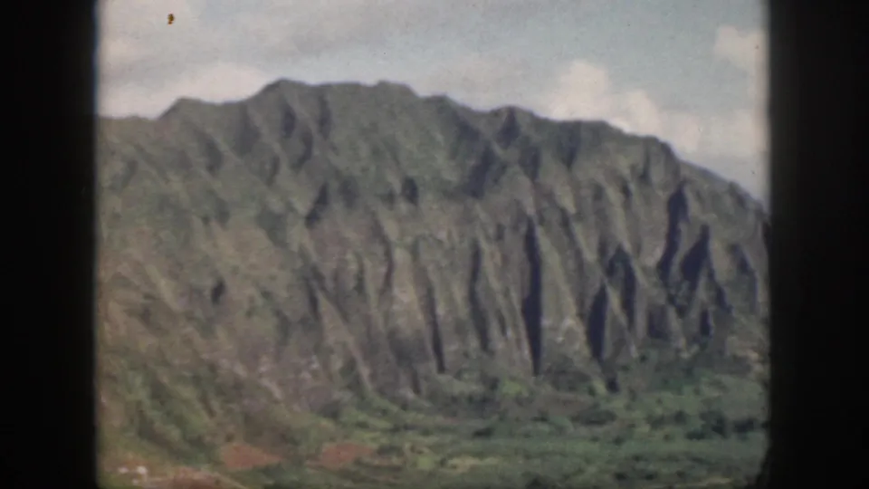 1959 Oahu 8mm frame of a wide green mountain wall with eroded vertical ridges and low valley vegetation.