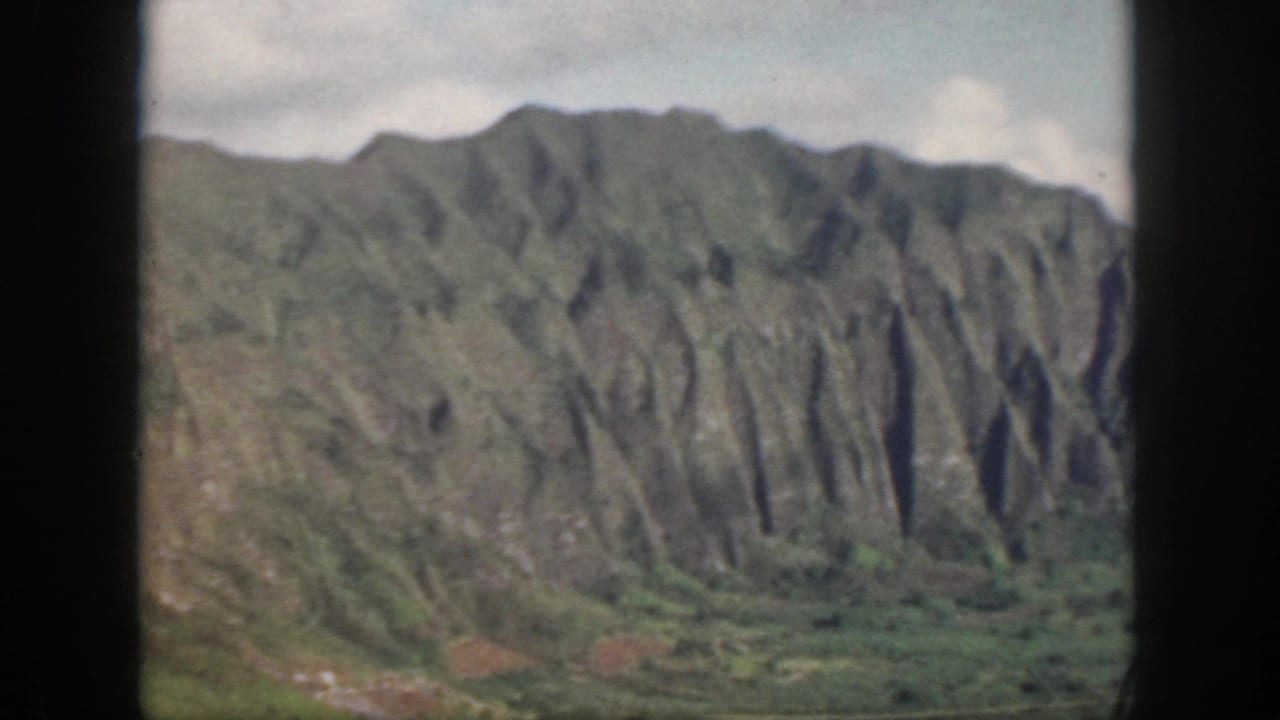 1959 Oahu home-movie frame showing verdant ridges and steep eroded mountain faces under cloud cover.