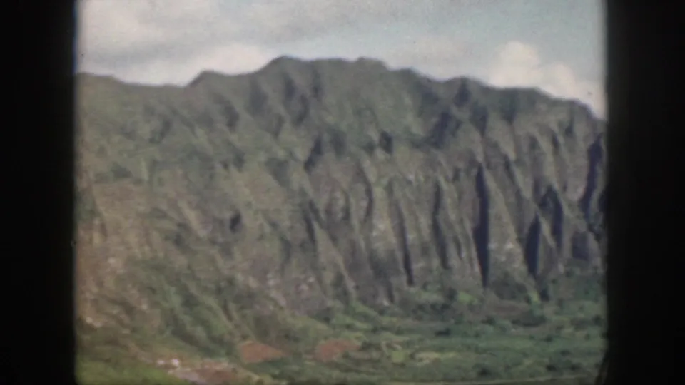 1959 Oahu home-movie frame showing verdant ridges and steep eroded mountain faces under cloud cover.