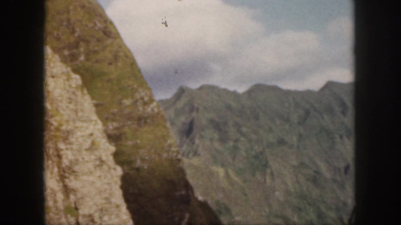 1959 Oahu home-movie frame showing a steep green ridge, pale rocky cliff face, distant mountains, and blue sky with clouds.