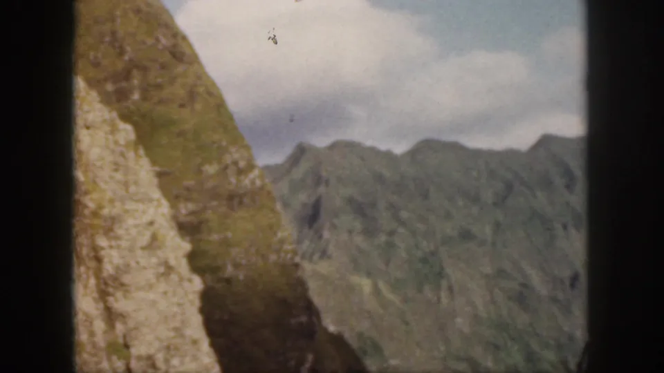 1959 Oahu home-movie frame showing a steep green ridge, pale rocky cliff face, distant mountains, and blue sky with clouds.