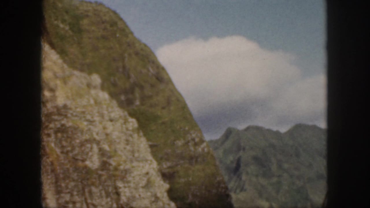 Rocky green hillside with distant ridges and clouded blue sky.