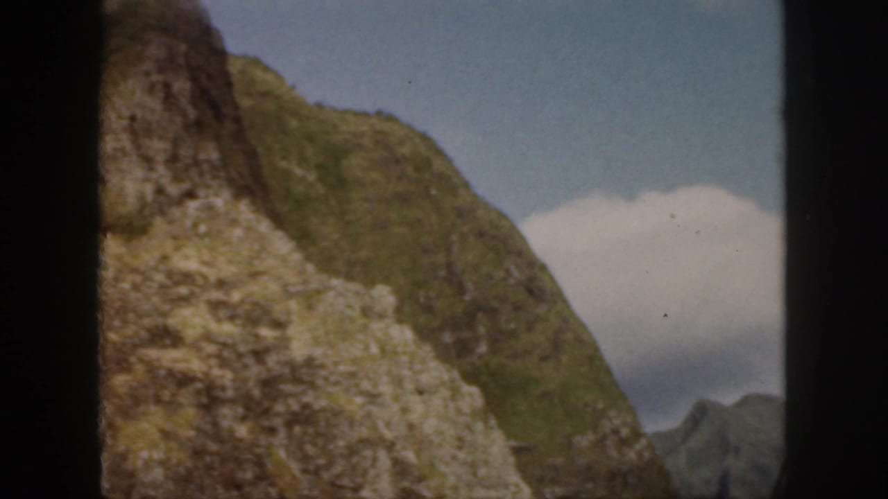 1959 Oahu home-movie view of a steep green slope with exposed pale rock and a small cloud against blue sky.