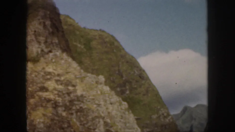 1959 Oahu home-movie view of a steep green slope with exposed pale rock and a small cloud against blue sky.