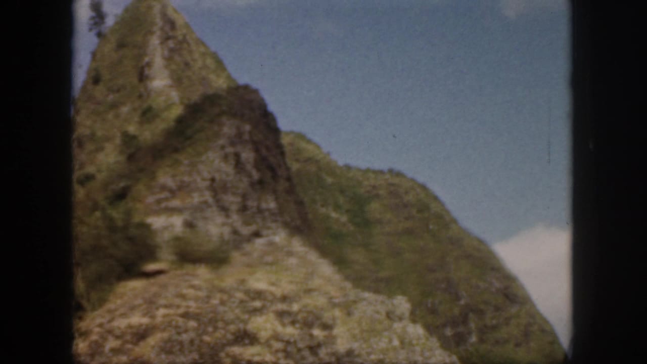 1959 Oahu 8mm frame showing a pointed rocky rise, green mountainside, and blue sky with a low white cloud near the edge.