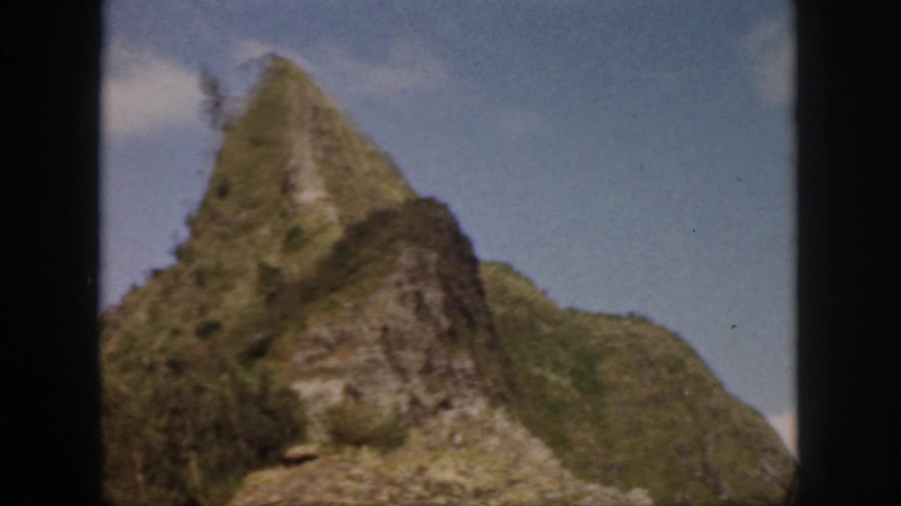 1959 Oahu 8mm frame showing a steep, foliage-covered cliff face with rocky ledges and blue sky.