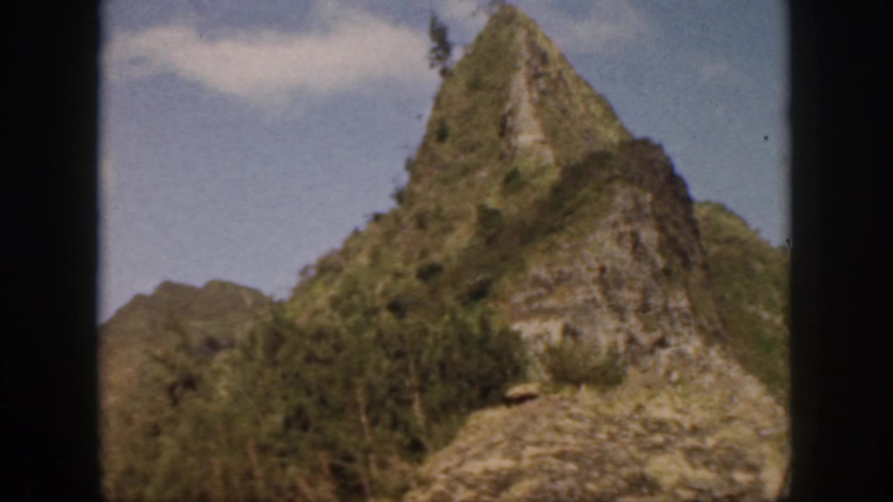 1959 Oahu frame with a rocky, vegetation-covered cliff peak and soft blue sky in the background.