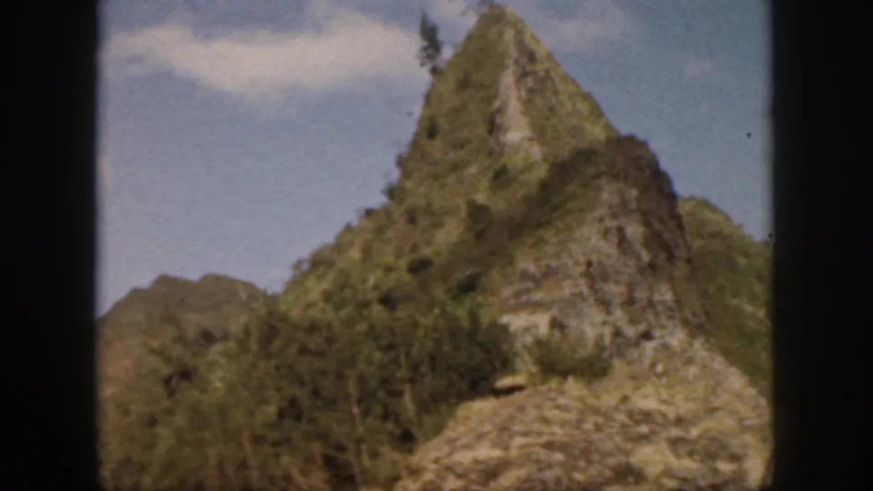 1959 Oahu frame with a rocky, vegetation-covered cliff peak and soft blue sky in the background.