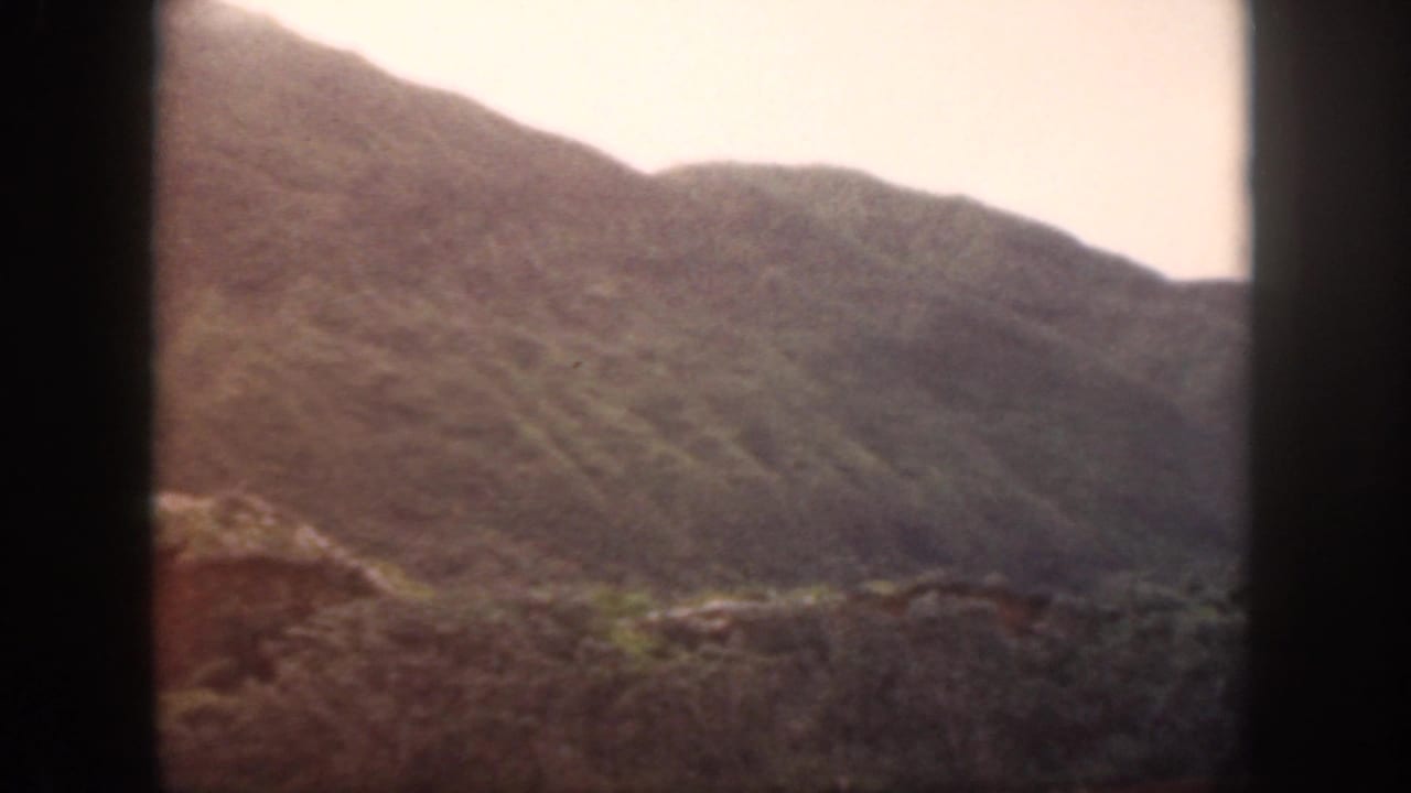 1959 Oahu 8mm frame showing a soft, distant mountain ridge with rocky foreground and pale daylight sky.