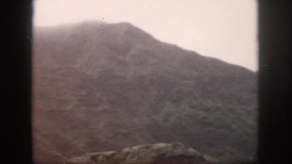 1959 Oahu 8mm frame showing a hazy mountain profile, muted hillside texture, and rocky foreground.