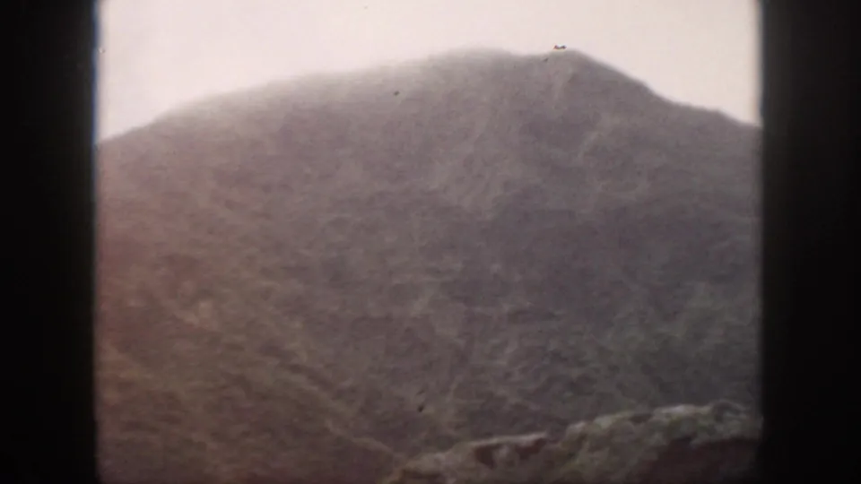 1959 Oahu frame showing a broad mountain slope, rocky lower edge, pale sky, and visible film mark near the upper frame.