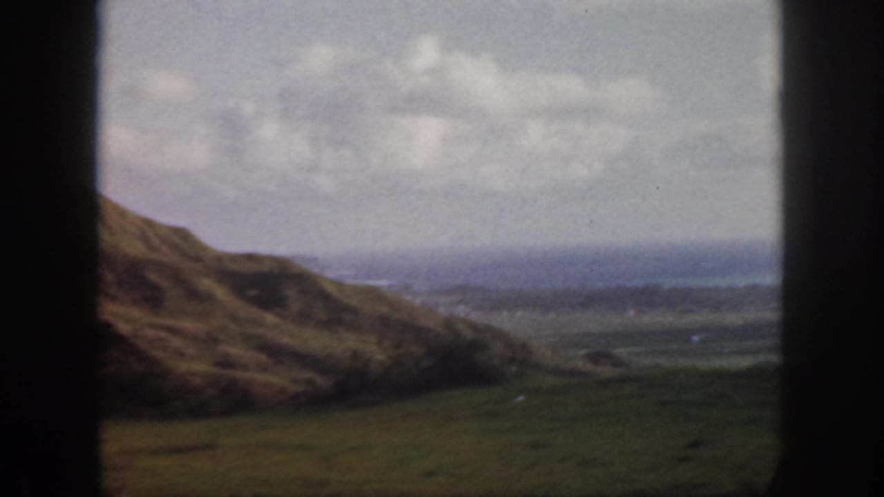 1959 Oahu home-movie frame showing grassy slopes, a broad valley, distant coast, cloudy sky, and dark window edges.