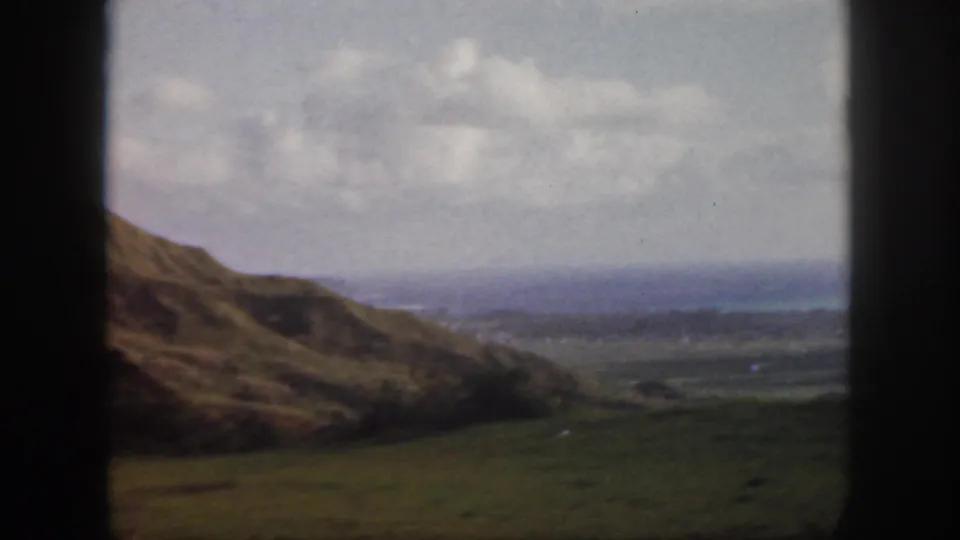 1959 Oahu home-movie frame showing grassy slopes, a broad valley, distant coast, cloudy sky, and dark window edges.