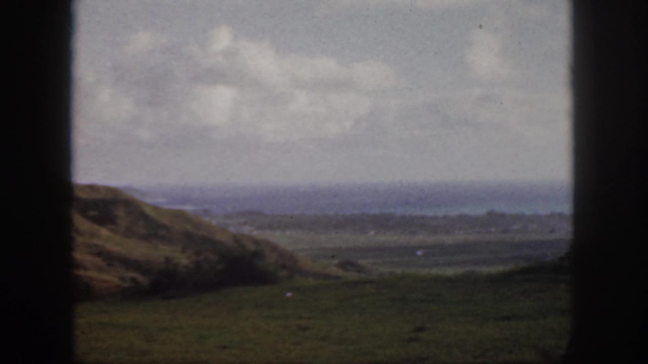 1959 Oahu 8mm frame with a rolling grassy slope, open valley floor, distant coastal water, cloudy sky, and window bars.
