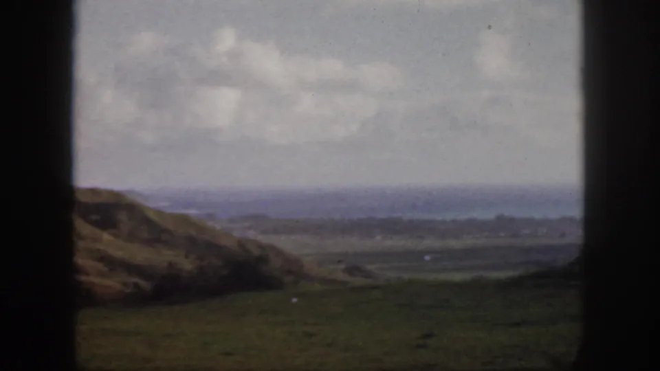 1959 Oahu 8mm frame with a rolling grassy slope, open valley floor, distant coastal water, cloudy sky, and window bars.