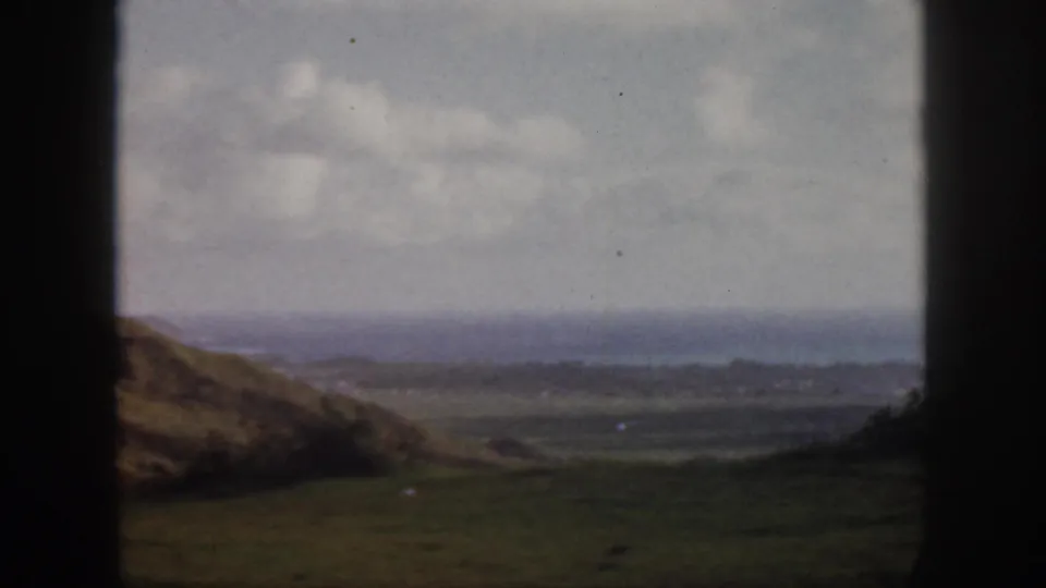1959 Oahu travel footage frame showing a muted green valley, hillside edge, distant coastline, cloudy sky, and heavy dark window borders.