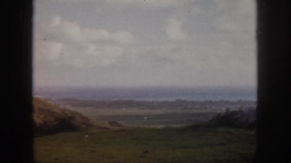 1959 Oahu 8mm frame looking across grassy foreground and valley toward a hazy coastline beneath a cloudy sky.