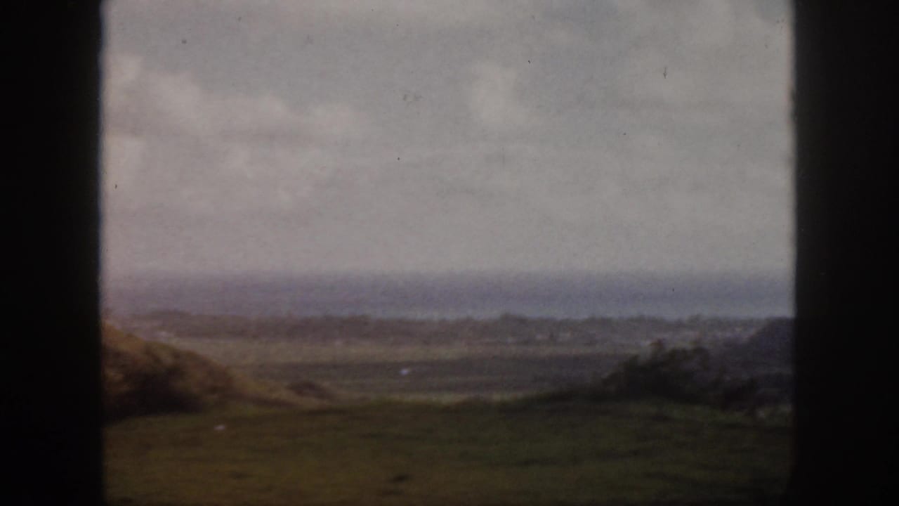 1959 Oahu 8mm frame showing a grassy overlook, distant shoreline, ocean haze, and cloudy sky with dark film-edge framing.