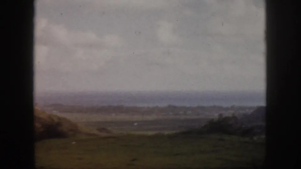 1959 Oahu 8mm frame showing a grassy overlook, distant shoreline, ocean haze, and cloudy sky with dark film-edge framing.