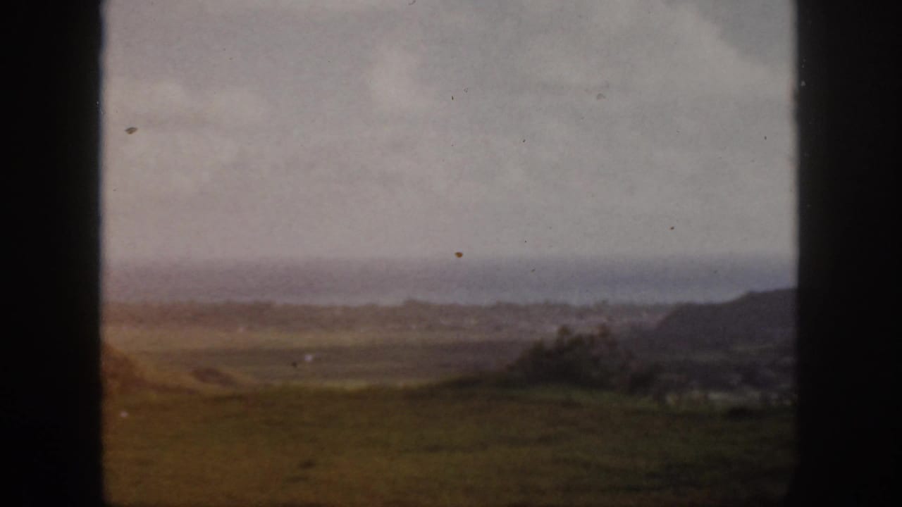 1959 Oahu home-movie view across open coastal grassland toward a hazy ocean horizon, partly bordered by dark frame edges.