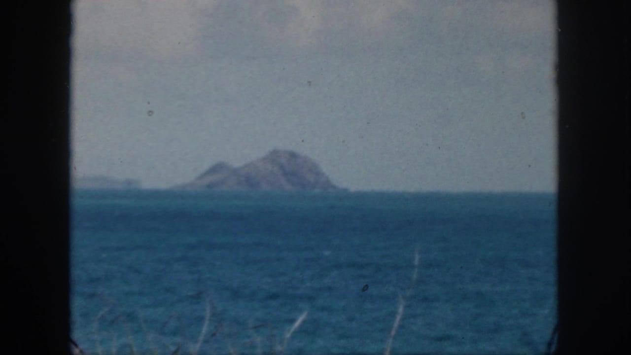 1959 Oahu 8mm frame showing a distant rocky islet beyond blue ocean with pale sky and dark film borders.