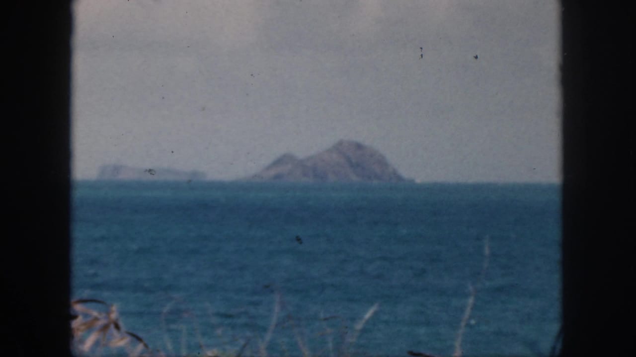 1959 Oahu coastal view with distant rocky islet, low offshore landforms, blue water, and faint vegetation near the frame edge.