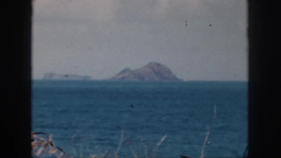 1959 Oahu coastal view with distant rocky islet, low offshore landforms, blue water, and faint vegetation near the frame edge.
