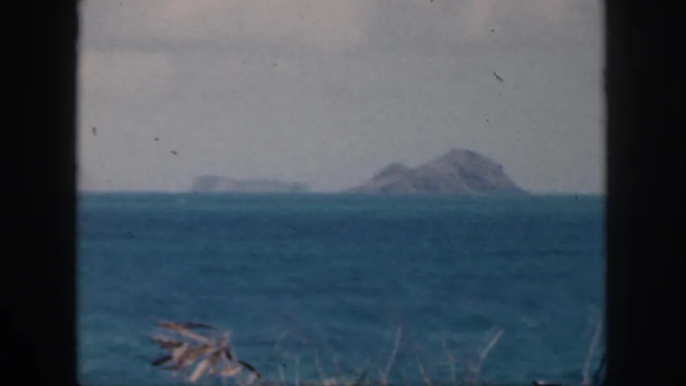 1959 Oahu 8mm frame with blue ocean, a distant rocky islet, pale sky, and sparse dry grasses along the lower edge.