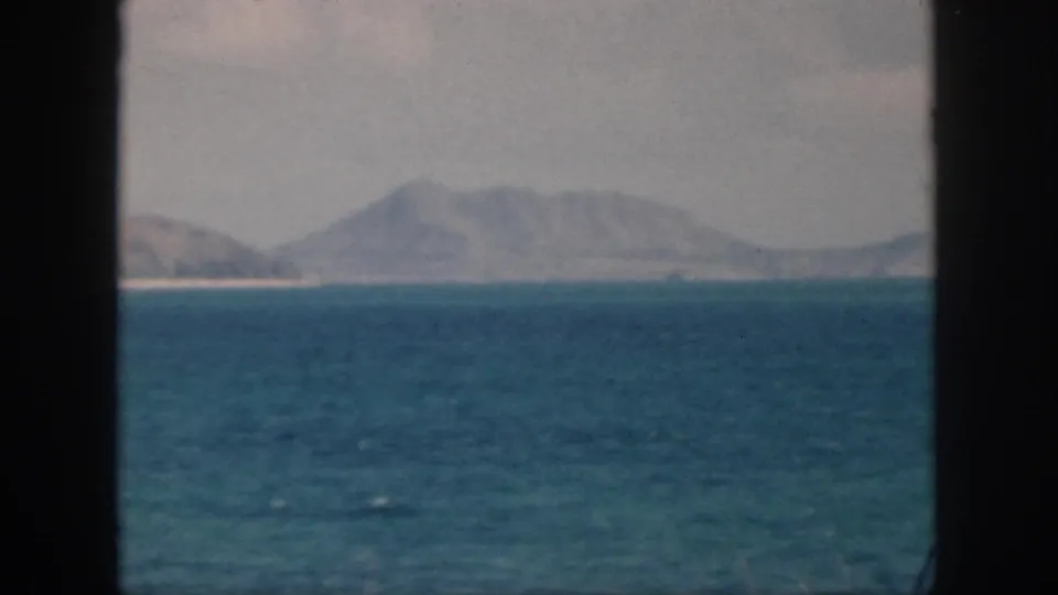1959 Oahu coastal landscape with turquoise water, a pale shoreline, and a broad hazy mountain in the distance.