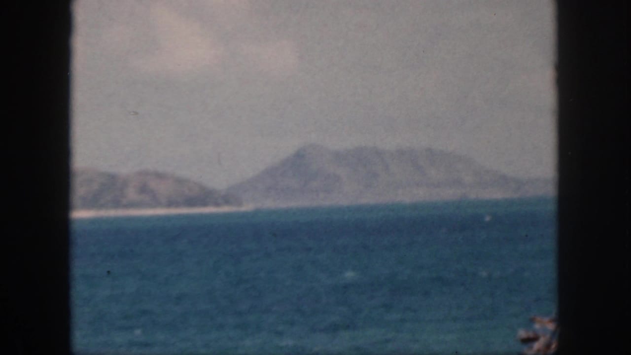 1959 Oahu home-movie frame showing blue water, distant hazy hills, and a bright shoreline through dark vertical borders.