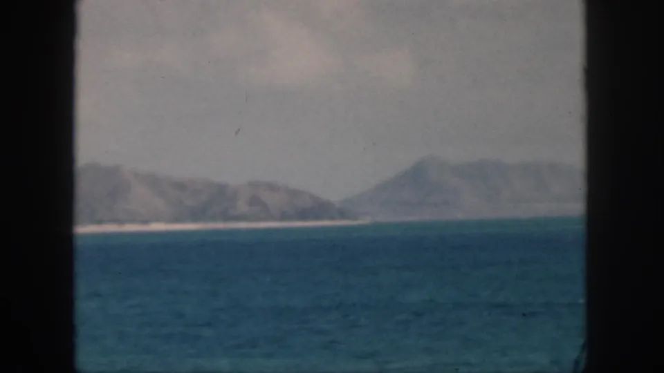 1959 Oahu travel footage frame with calm blue ocean, hazy coastal hills, pale beach line, and dark borders at the sides.
