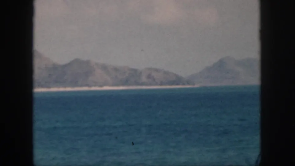 1959 Oahu home-movie frame showing a hazy ridge, pale shore, and blue ocean under a bright sky with film softness.
