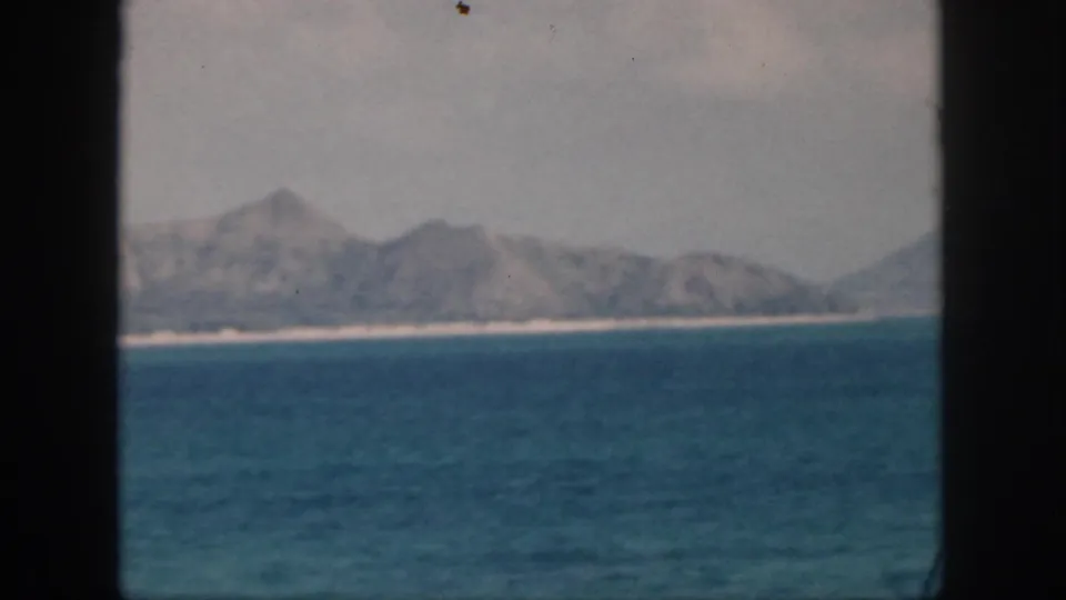 1959 Oahu frame with blue ocean in the foreground, distant hazy hills, and a pale coastal strip in soft 8mm color.